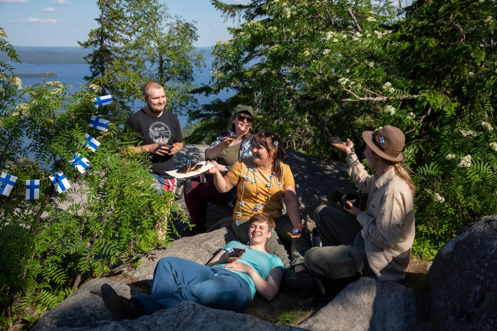 Kuvassa on viiden hengen seurue, joka on kokoontunut luonnon keskelle juhlimaan. Taustalla avautuu näköala ja sininen järvimaisema, ja seuruetta ympäröivät vihreät puut ja pensaat, joita on koristeltu pienillä Suomen lipuilla.