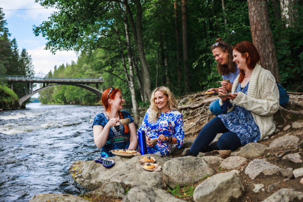 Kuvassa neljä naista syö eväitä kosken rannalla. Naiset hymyilevät toisilleen. Taustalla näkyy kosken ylittävä silta ja kesäistä metsämaisemaa.