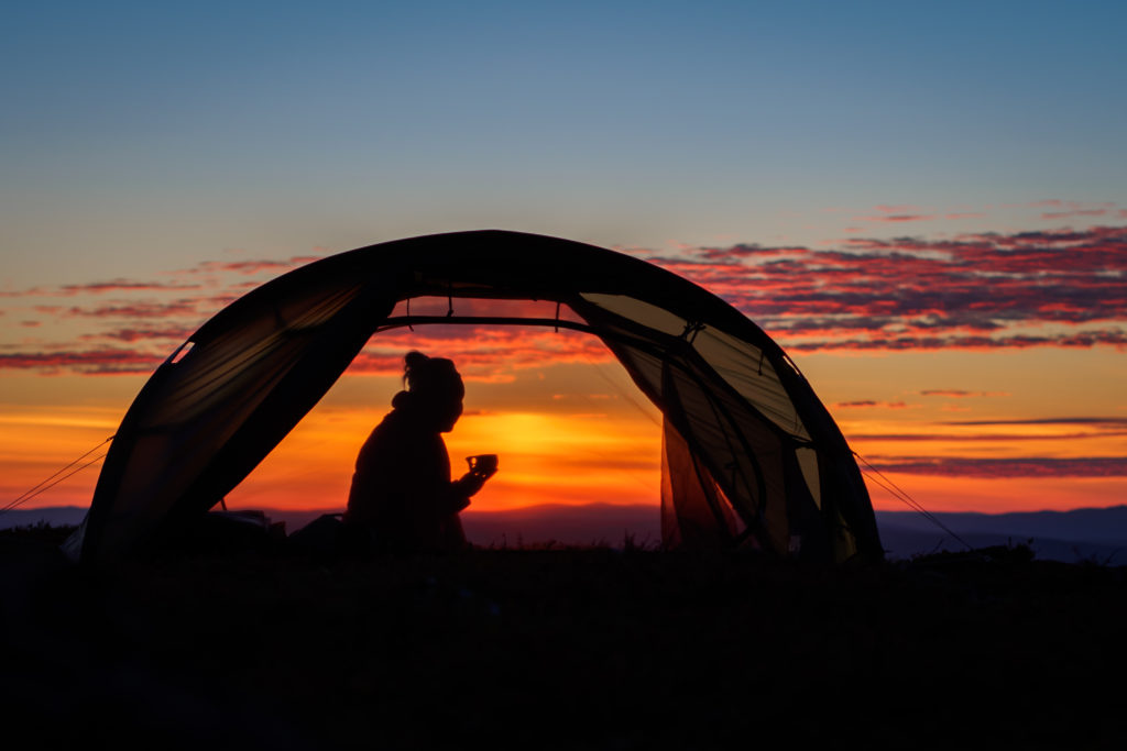 Kuvassa näkyy auringonlaskua vasten teltassa istuva nainen. Hänen voi nähdä hymyilevän ja pitelevän kädessään kuksaa. Tärkeä osa Suomen luonnon päivää on ulkona nukkuminen.