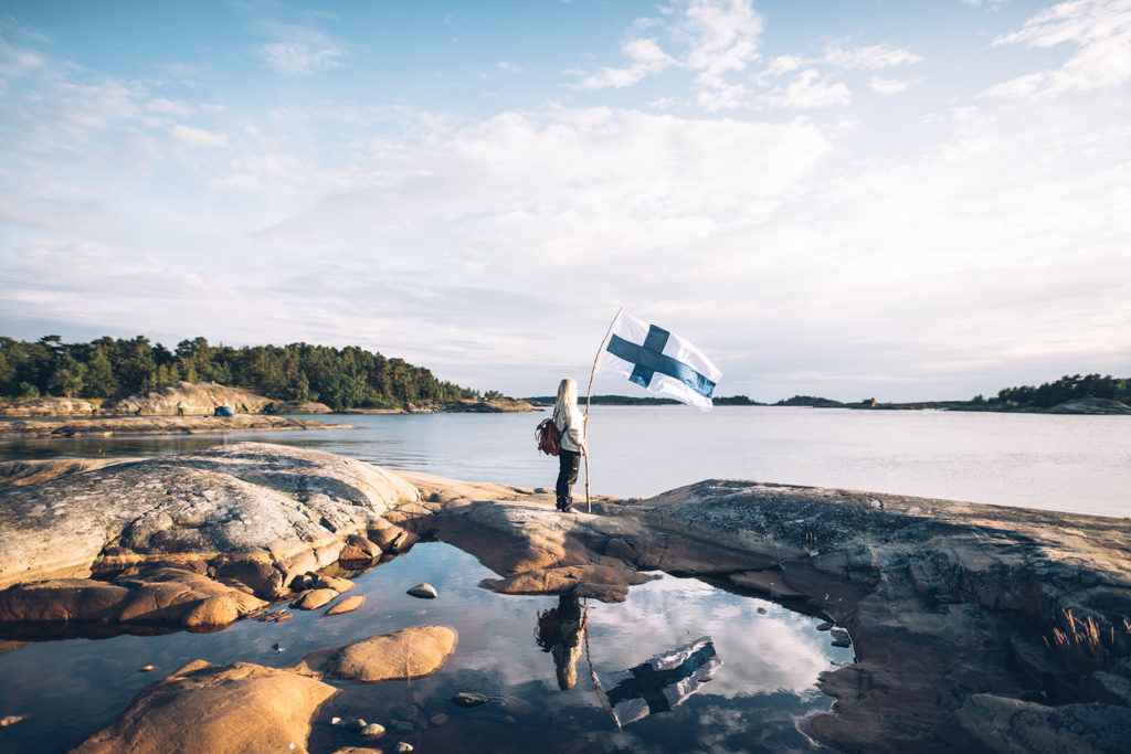 Henkilö pitämässä Suomen lippua rantakalliolla veden ääressä